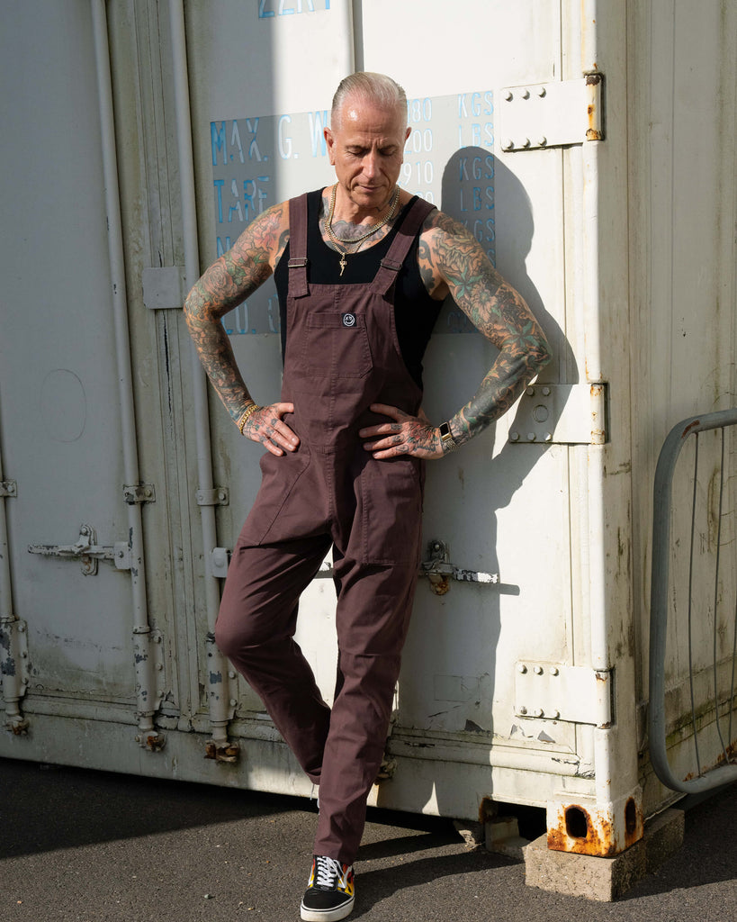 James with tattoo sleeves wears Rebel Romance Plum Mocha Stretch Twill Dungarees over a black vest, standing in sunlight against a white shipping container.