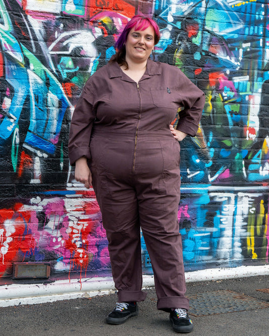 Chloe models the Plum Mocha Boiler Suit by Rebel Romance, styled with rolled cuffs and casual trainers. The utility jumpsuit features a zip front, collar and side pockets, photographed against a vibrant street art wall.