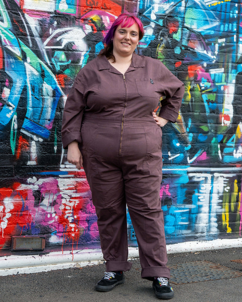 Chloe models the Plum Mocha Boiler Suit by Rebel Romance, styled with rolled cuffs and casual trainers. The utility jumpsuit features a zip front, collar and side pockets, photographed against a vibrant street art wall.
