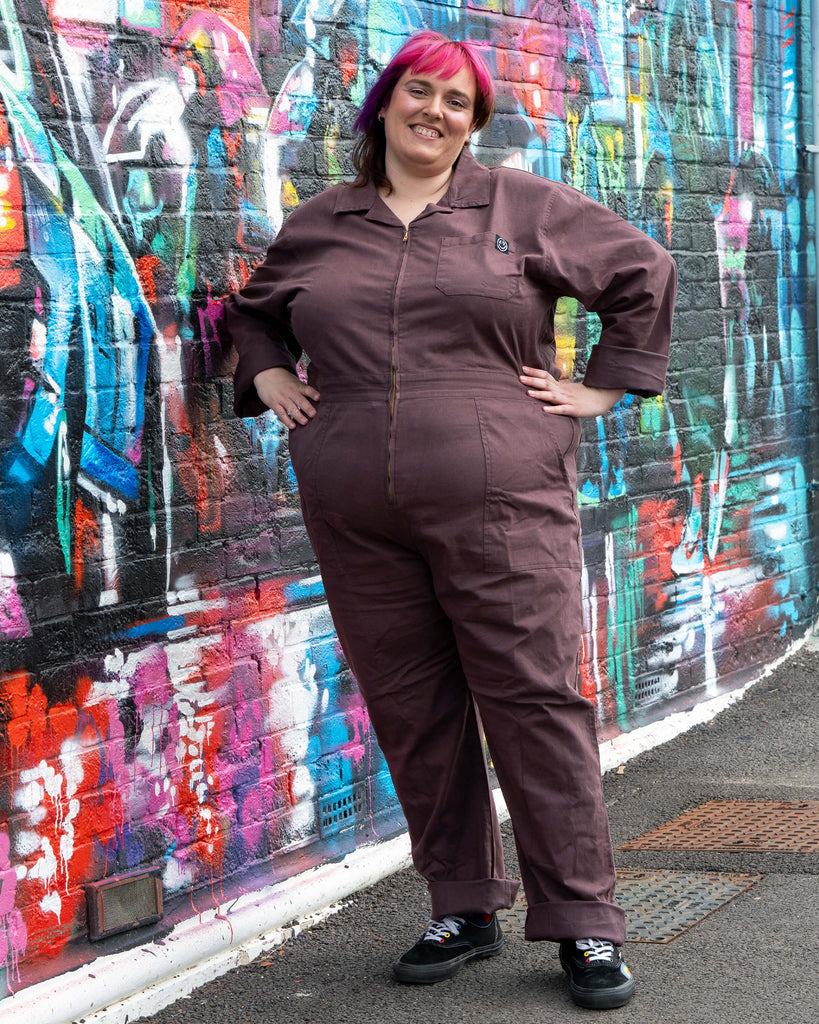 Chloe wears the Rebel Romance Plum Mocha Boiler Suit with hands on hips, styled with black trainers. The jumpsuit has a zip fastening, collar and functional pockets, photographed outdoors with a colourful graffiti mural backdrop.