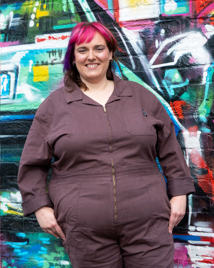 Portrait of Chloe smiling in the Plum Mocha Boiler Suit by Rebel Romance. The zip-up jumpsuit with logo chest patch is styled for comfort and edgy everyday wear, shot against a brightly painted graffiti wall.