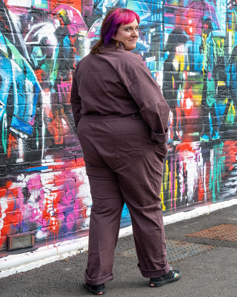 Chloe models the Plum Mocha Boiler Suit by Rebel Romance, styled with rolled cuffs and casual trainers. The utility jumpsuit features a zip front, collar and side pockets, photographed against a vibrant street art wall.
