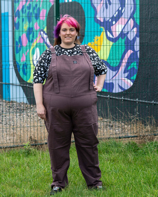 Full-length shot of Chloe in the Plum Mocha Dungarees by Rebel Romance. Styled with a ghost-print T-shirt and photographed outdoors in front of a mural, these dungarees showcase an edgy, playful alternative style.