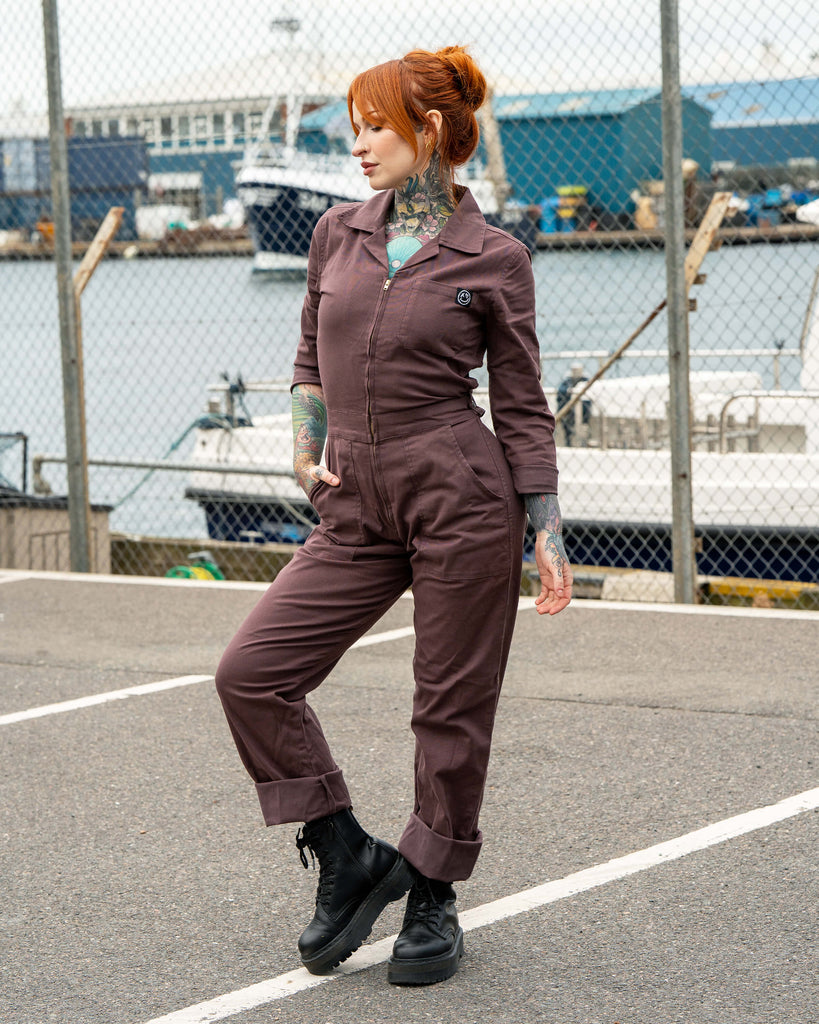 Erin poses side-on wearing the Rebel Romance Plum Mocha Boiler Suit, styled with rolled cuffs and chunky black lace-up boots. The utility jumpsuit has a zip front, collar and pockets, photographed by a harbour with boats in the background.