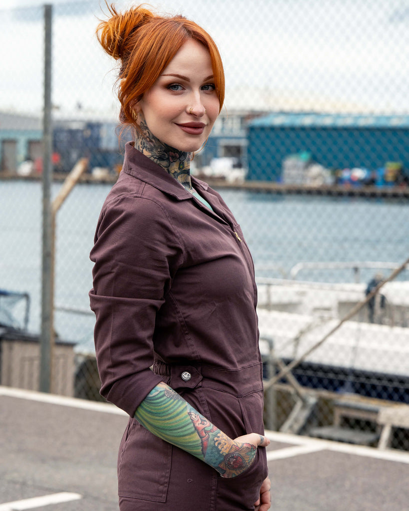 Side view of Erin wearing the Plum Mocha Boiler Suit by Rebel Romance. The fitted jumpsuit has a zip front, collar and pockets, styled with rolled cuffs and chunky black boots, shot by the waterfront with boats in the background.