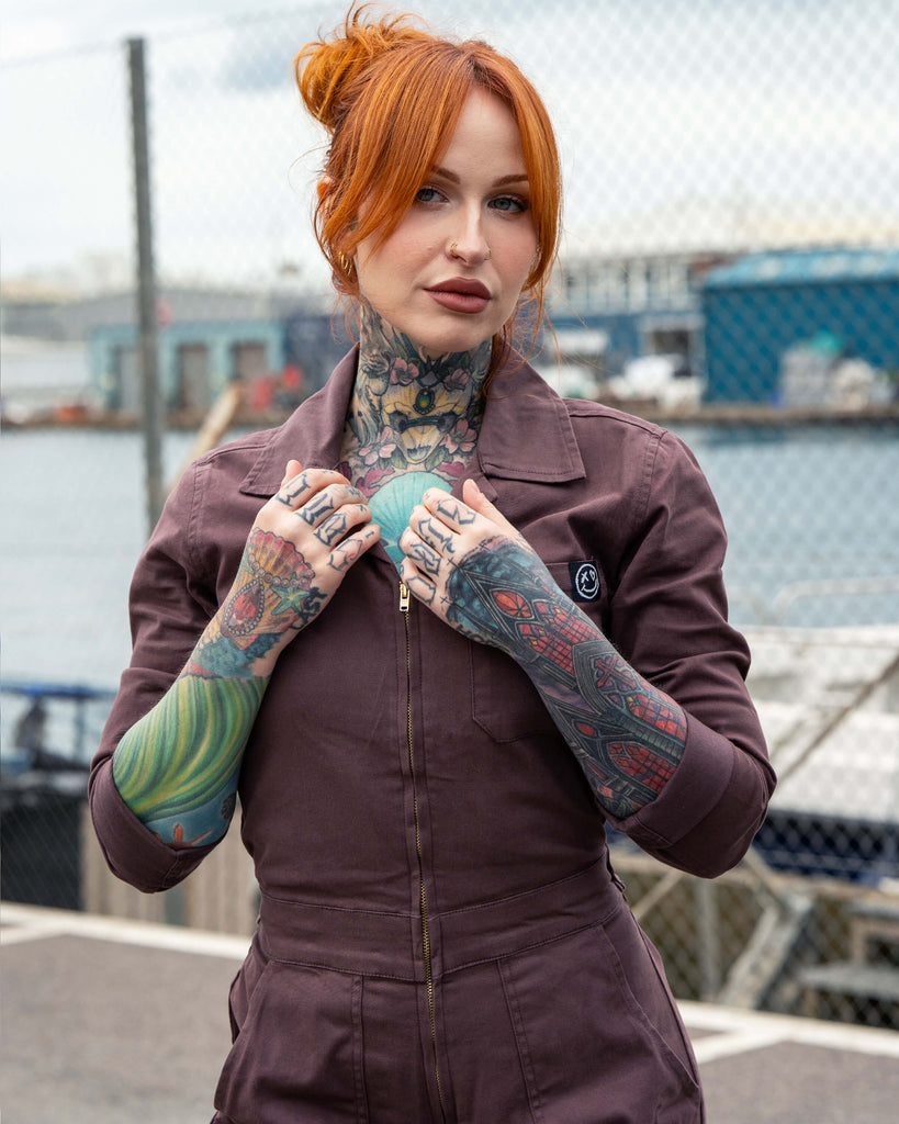 Erin wears the Plum Mocha Boiler Suit by Rebel Romance in a close-up shot. The zip front and collar are visible, styled with bold tattoos and rings, giving an edgy alternative vibe perfect for slow fashion streetwear.