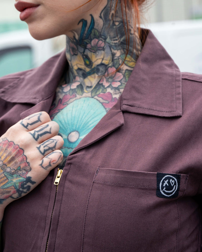 Close up of Erin in the Plum Mocha boiler suit with a tattooed hand in the shot holding the collar. The happy face graffiti style logo is on the front pocket.