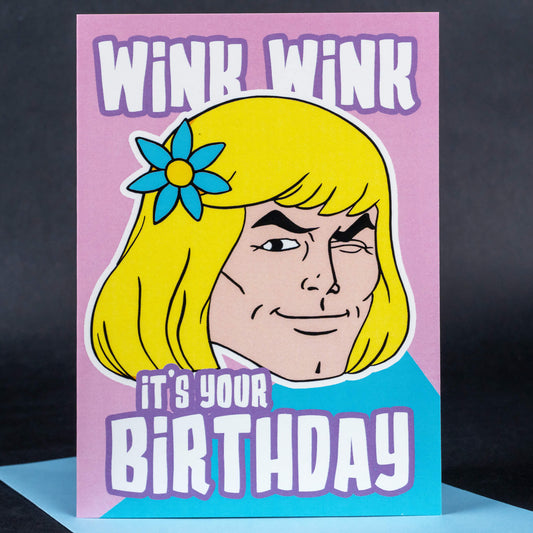 Humorous birthday card with a retro-style illustration of a winking man in blonde hair and a flower accessory, accompanied by the text ‘WINK WINK, it’s your BIRTHDAY’ in bold pastel colours.