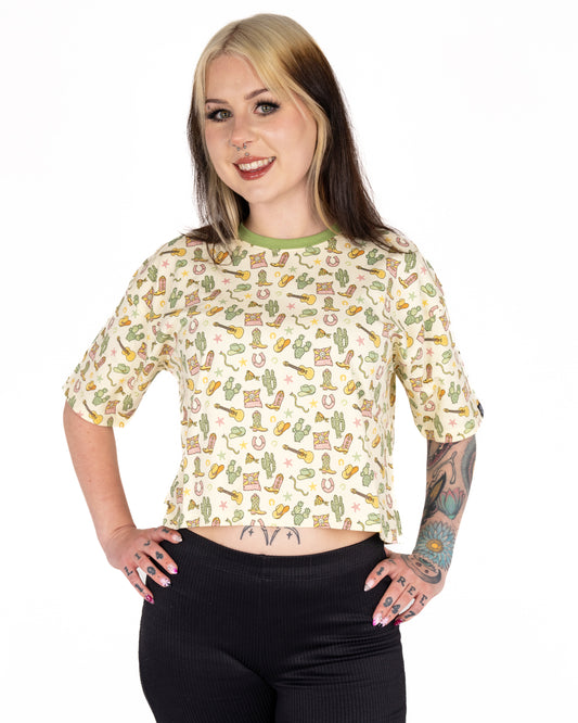 Florence (@florencerosaria) models the Rebel Romance x Yasmin Cousins Cowboy Cropped Boxy Tee with hands on hips. The cream tee has a green neckline and playful Western print. Florence wears a size 10 and is 5'3", styled with high-waisted black trousers. Rebel Romance is a small UK brand celebrating bold, ethical fashion.