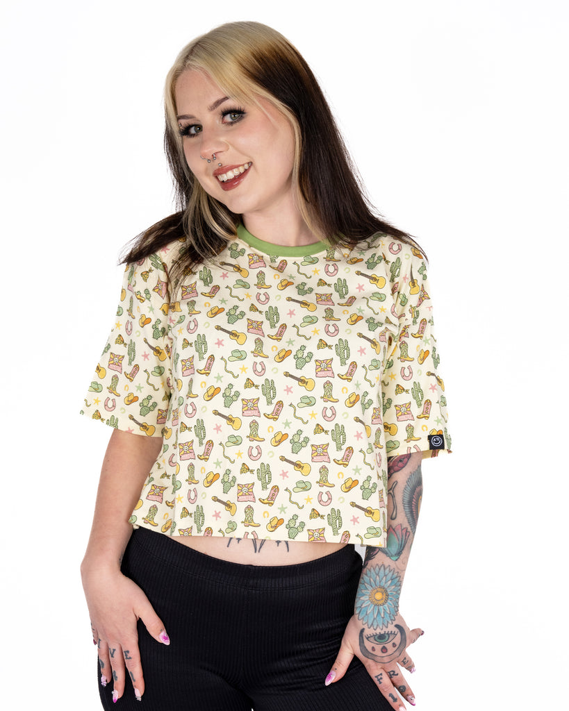 Florence (@florencerosaria) smiles wearing the Rebel Romance x Yasmin Cousins Cowboy Cropped Boxy Tee in size 10. The cream boxy tee features an all-over print of boots, cacti, and hats. Rebel Romance is a UK-based brand known for quirky, ethical fashion celebrating alternative aesthetics.