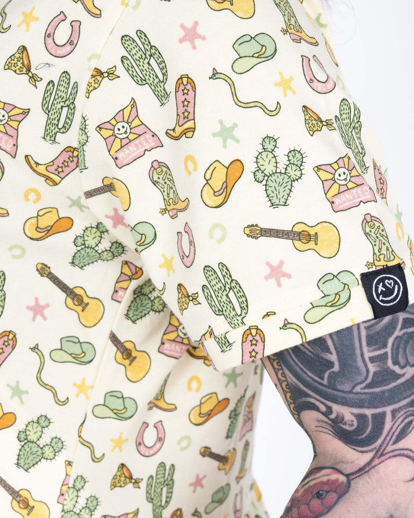 Close-up of sleeve on the Rebel Romance cowboy smock dress showing cacti, cowboy hats, guitars, snakes, and a pink “WANTED” poster motif.