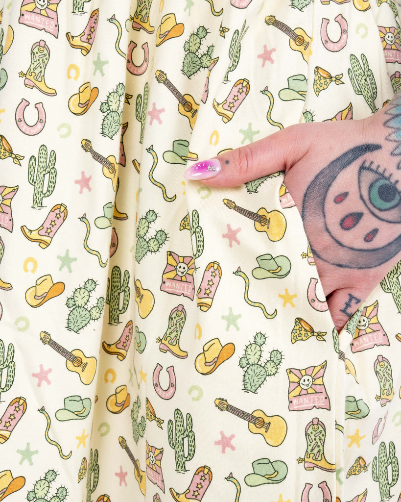 Close-up of the skirt of the cream cowboy print dress with Florence’s hand in the pocket, showing yellow guitars, boots, and green hats.