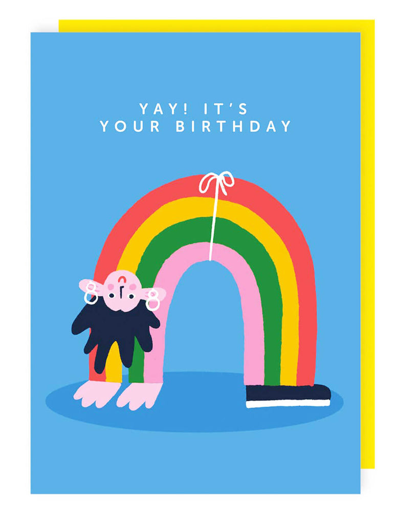 A bright blue birthday card featuring a rainbow character with pink arms, a black beard, hoop earrings, and one black sneaker. The words “YAY! IT’S YOUR BIRTHDAY” are printed in white above the rainbow, with a yellow envelope peeking out behind the card.
