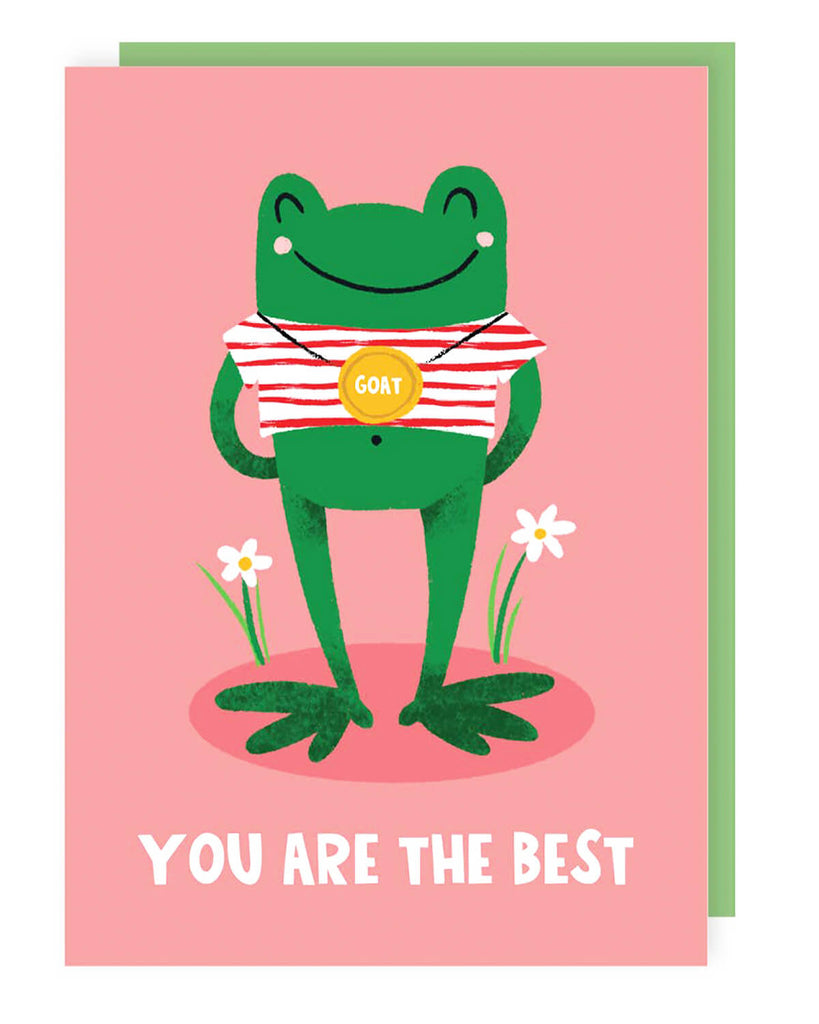 A pink greeting card featuring a happy green frog in a red and white striped top, wearing a gold medal that says “GOAT” around its neck. The card has “YOU ARE THE BEST” written in bold white letters at the bottom, with two small daisies growing beside the frog. A green envelope peeks out from behind the card.
