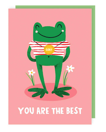 A pink greeting card featuring a happy green frog in a red and white striped top, wearing a gold medal that says “GOAT” around its neck. The card has “YOU ARE THE BEST” written in bold white letters at the bottom, with two small daisies growing beside the frog. A green envelope peeks out from behind the card.
