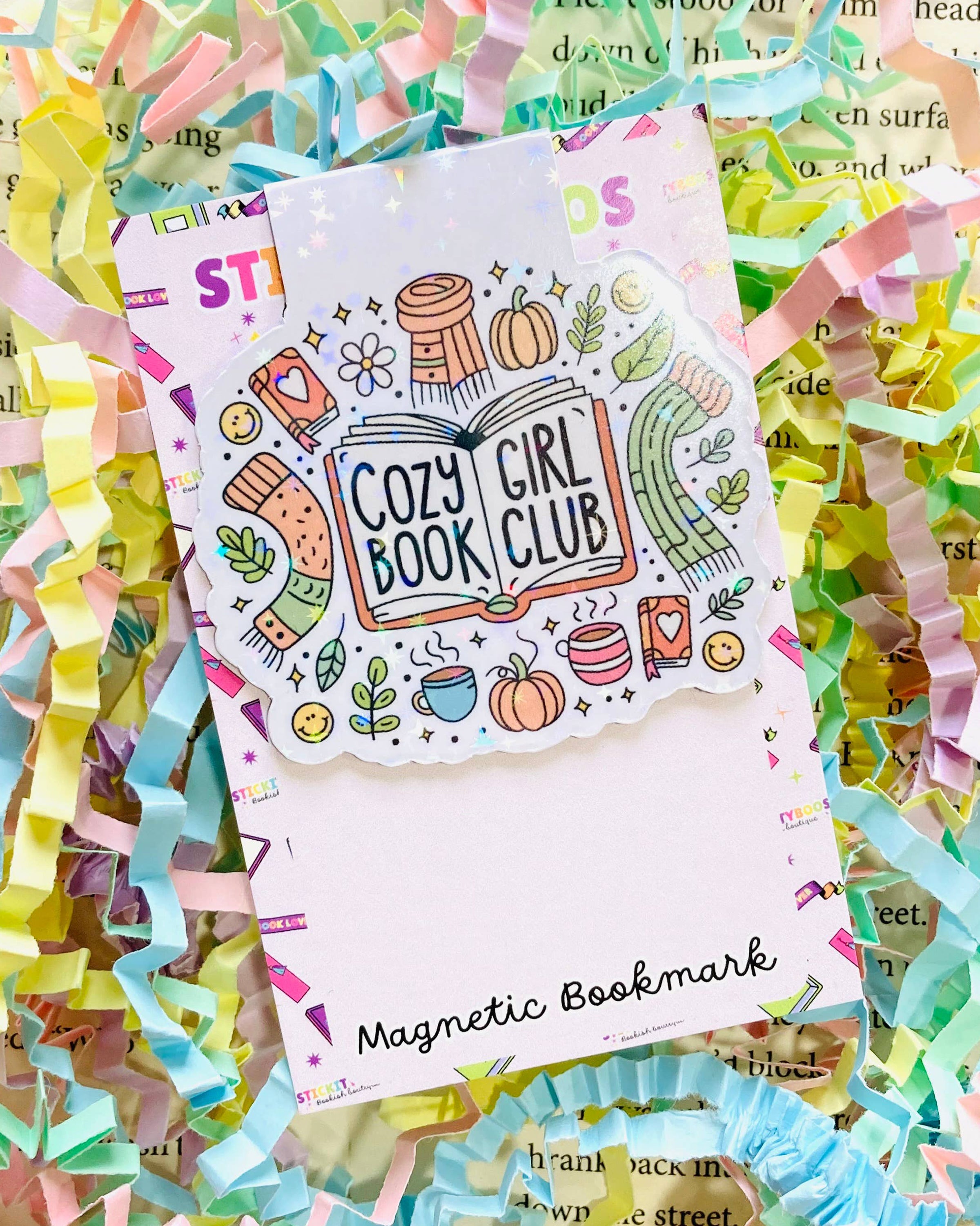 A Rebel Romance magnetic bookmark by The Stickityboo Co with a cute “Cozy Girl Book Club” design. Surrounded by pastel shredded paper, the illustrated bookmark features pumpkins, scarves, mugs, and plants, celebrating cosy autumn vibes and book lovers.