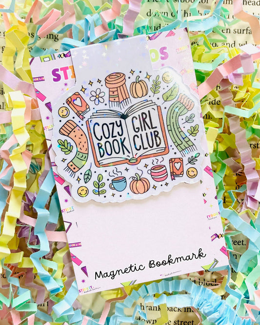 A Rebel Romance magnetic bookmark by The Stickityboo Co with a cute “Cozy Girl Book Club” design. Surrounded by pastel shredded paper, the illustrated bookmark features pumpkins, scarves, mugs, and plants, celebrating cosy autumn vibes and book lovers.