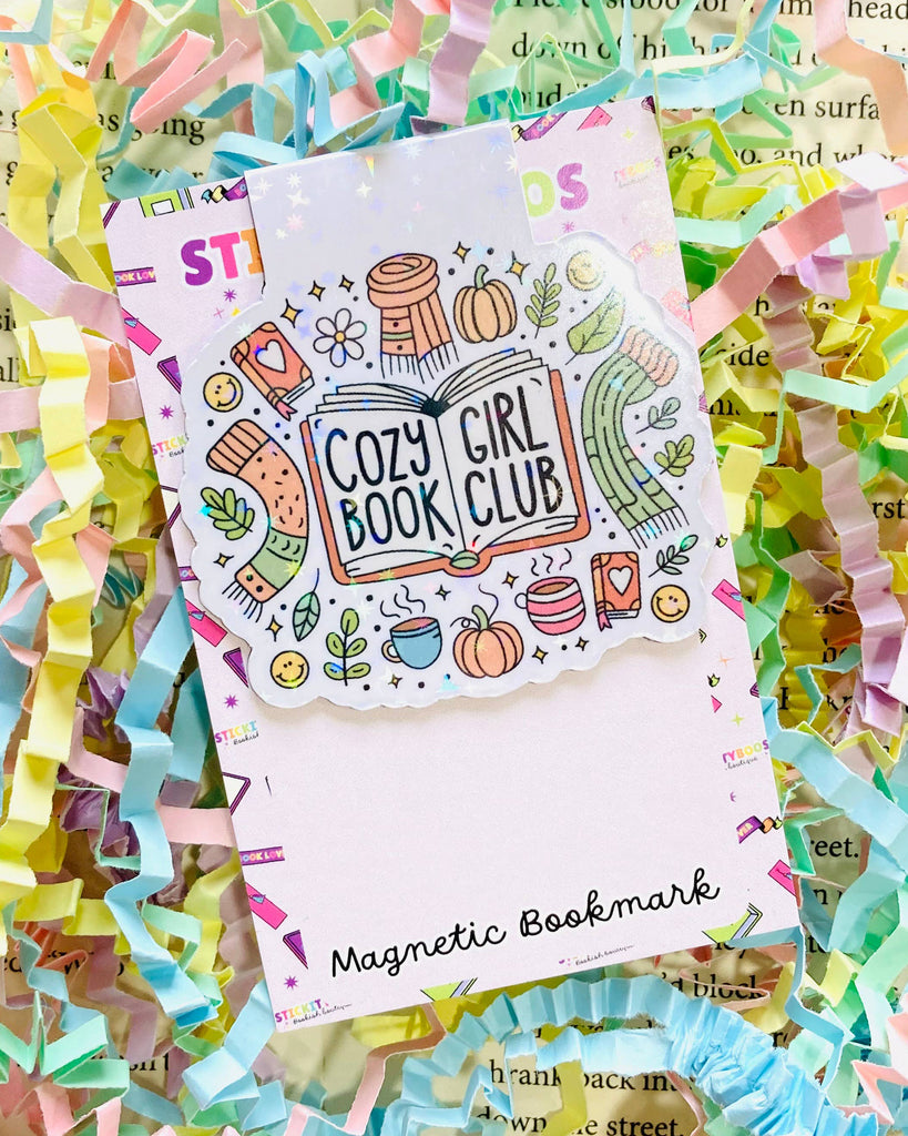 A Rebel Romance magnetic bookmark by The Stickityboo Co with a cute “Cozy Girl Book Club” design. Surrounded by pastel shredded paper, the illustrated bookmark features pumpkins, scarves, mugs, and plants, celebrating cosy autumn vibes and book lovers.