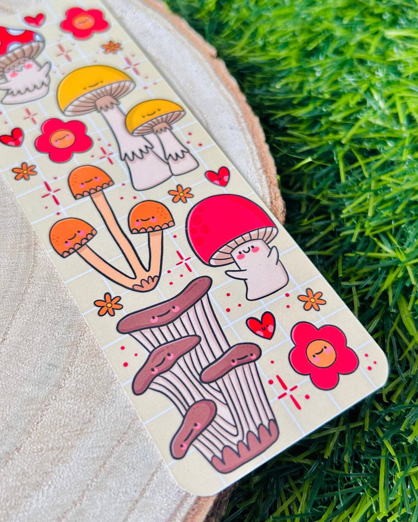 Flat lay of the Fungi Cuties Bookmark by Kelly Lou on a wooden surface and grass. Features happy mushrooms, flowers and hearts on a soft beige grid background. A cute and unique gift from Rebel Romance, a UK alternative lifestyle brand.