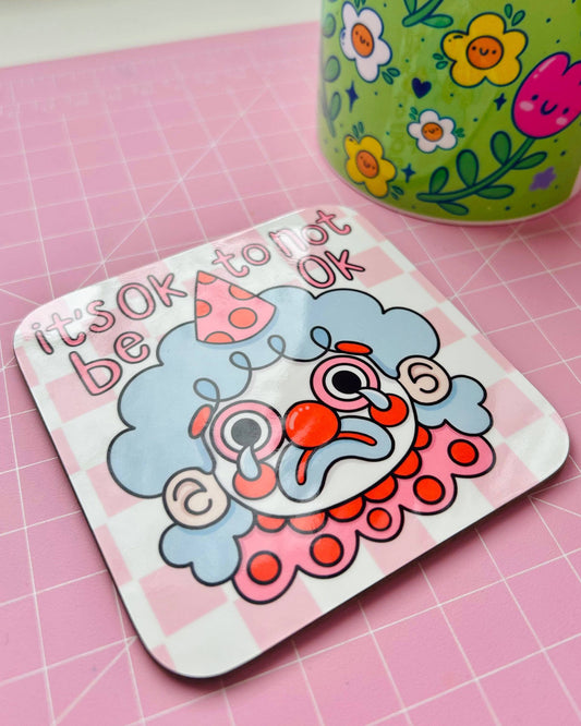 Glossy square coaster from Rebel Romance featuring a crying cartoon clown with blue hair and a pink party hat, surrounded by the words "it's OK to not be OK" in playful pink font. The coaster rests on a pink cutting mat next to a floral mug. This colourful coaster is designed by Kellylou and celebrates emotional honesty with quirky style.