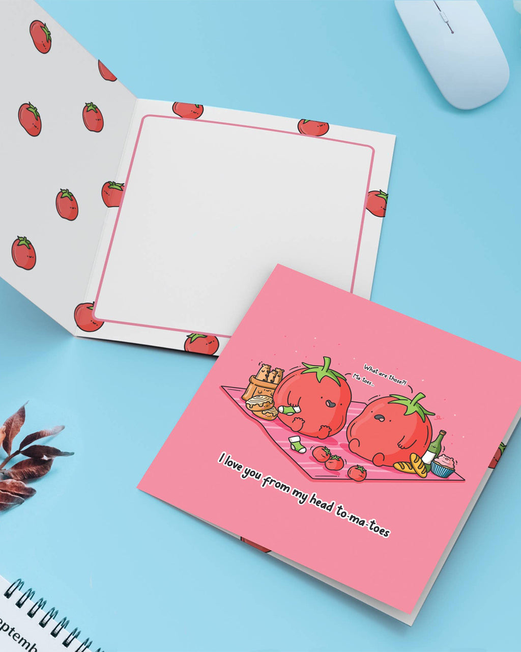 Open greeting card lying on a light blue surface. Inside is decorated with scattered tomato illustrations, with writing space outlined in pink. Beside it lies the closed card showing the tomato pun design. Sold by Rebel Romance in the UK.