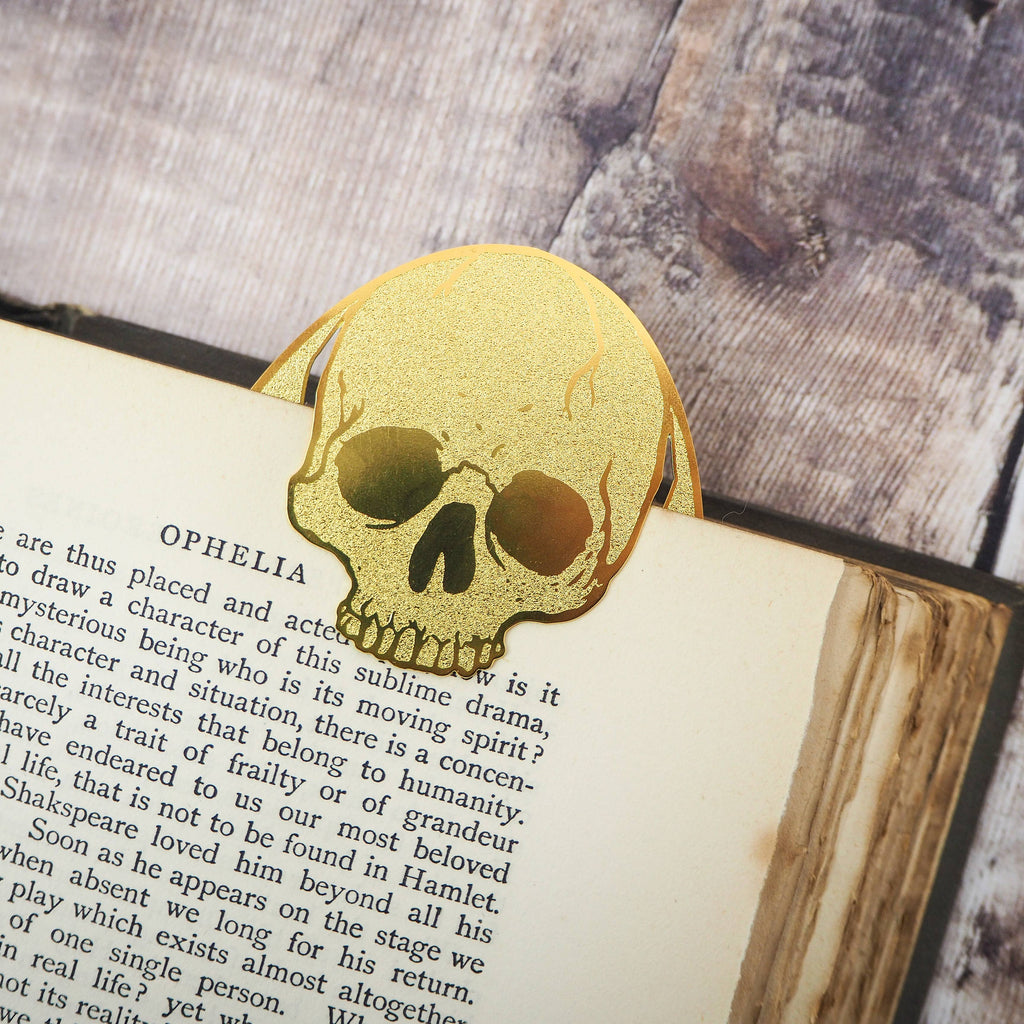 A brass bookmark featuring an oval shape with a skull design and floral etchings. The engraving reads, “To read, or not to read, that is the question,” inspired by Shakespeare’s Hamlet. The bookmark is placed inside an open vintage book on a wooden surface.