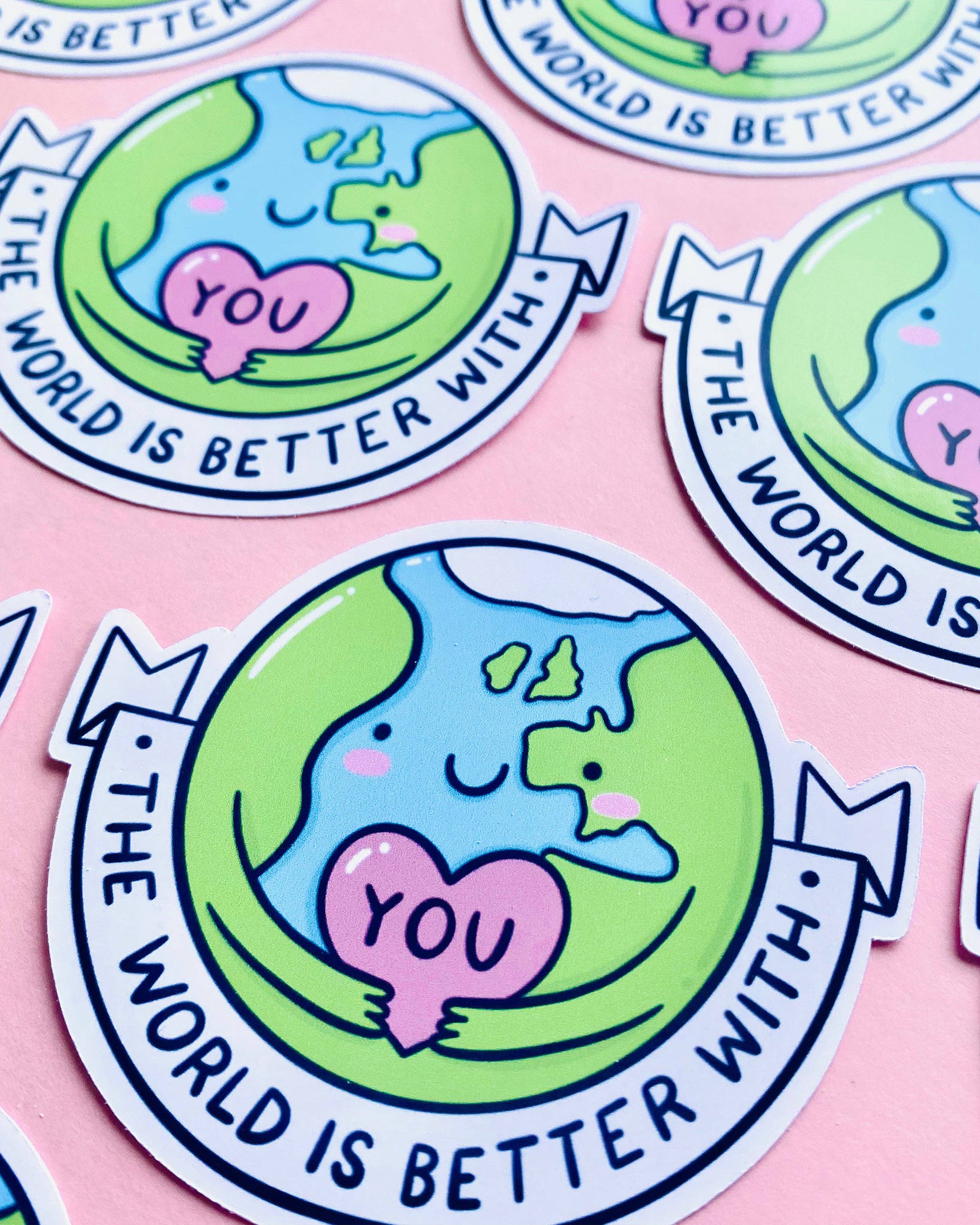 Flat-lay of several “The World Is Better With You” stickers from Rebel Romance. The smiling Earth design, illustrated by Kellylou, holds a pink heart and is surrounded by a motivational message. Set against a pastel pink background, perfect for mental health positivity and journalling.