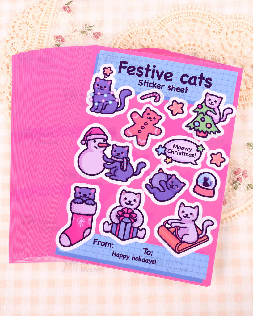 A stack of Christmas Festive Cats Sticker Sheets by Rebel Romance displayed on a soft cream lace background. The top A5 sheet shows adorable Mona Mauve illustrated cats celebrating the winter season including a snowman scene, a Christmas tree cat, a gingerbread cat and a cat in a stocking. The bright pink sheet adds a fun and festive feel, ideal for decorating gifts, journals and stationery.