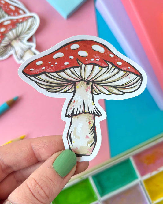 A close-up of a hand holding a red toadstool mushroom sticker with a white stem and whimsical details. The vinyl sticker is professionally printed and water-resistant. Designed by Stacey McEvoy Caunt and sold by Rebel Romance, a UK-based indie brand embracing slow fashion, alternative style and magical gifts.