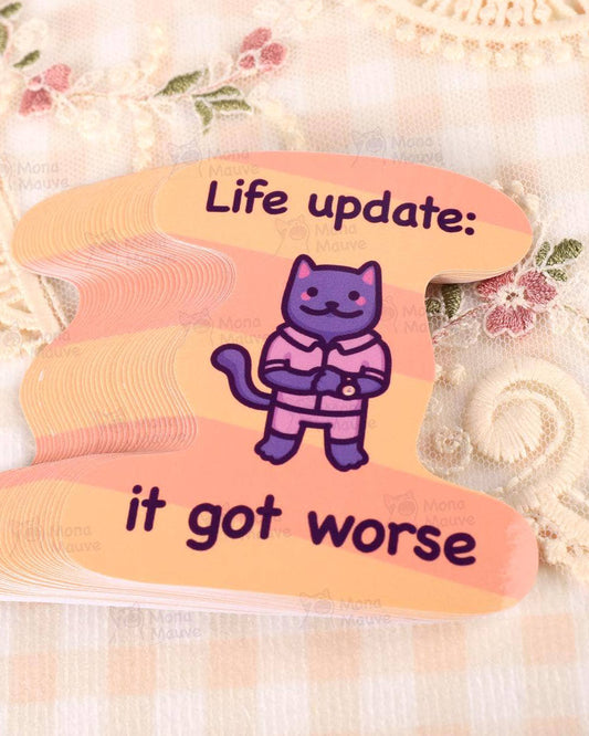 Close-up of Rebel Romance’s funny cat meme vinyl sticker by Mona Mauve, featuring a purple cartoon cat in pink pyjamas with the caption “Life update: it got worse.” Set against a peachy striped background with vintage-style lace and floral fabric underneath. This waterproof sticker is perfect for laptops, journals, or as a relatable gift for millennials with a dark sense of humour.