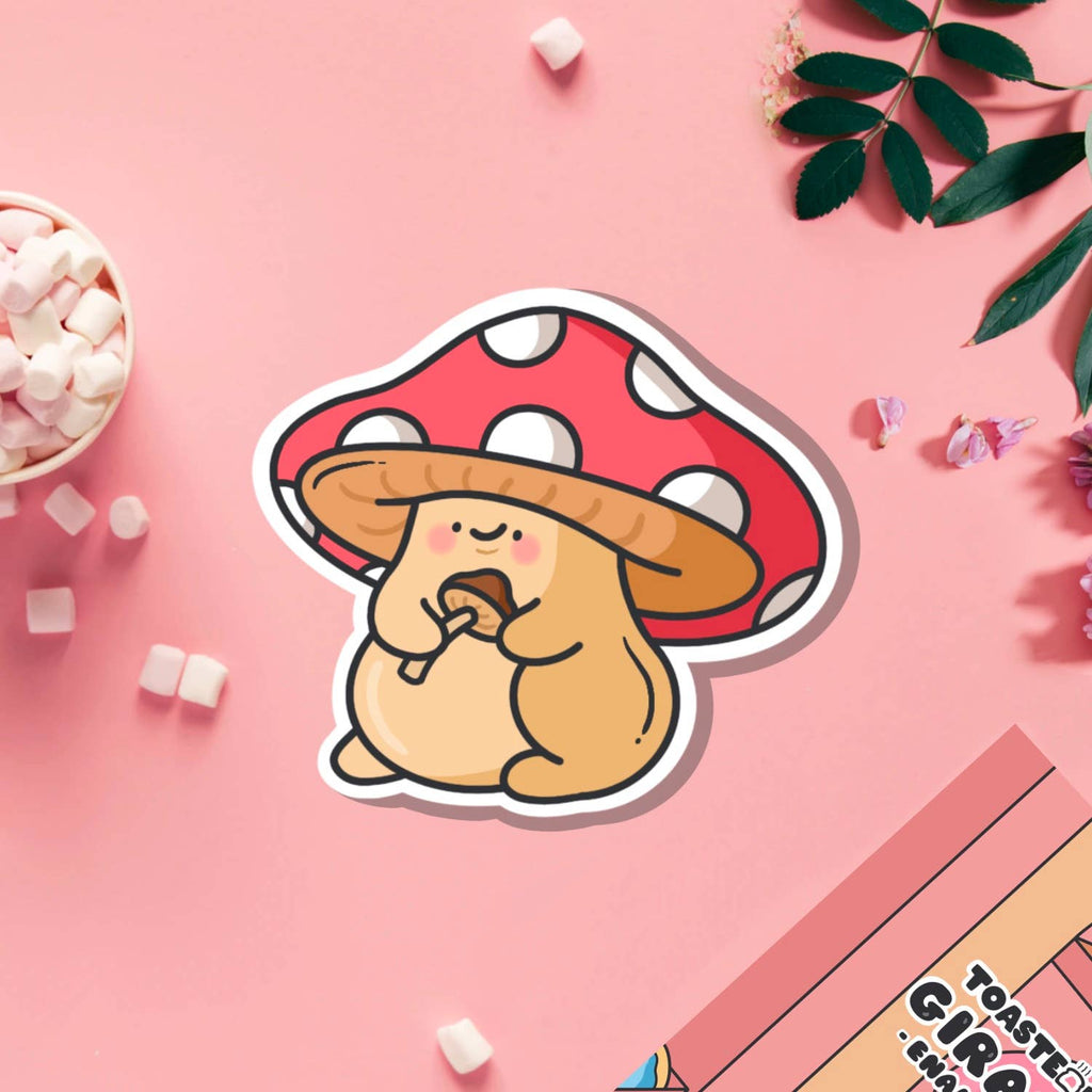 A cute cartoon-style vinyl sticker of a chubby tan mushroom character with a red and white spotted cap, holding a smaller mushroom in its hands. Displayed on a blue background beside a teacup and flower. Waterproof, glossy finish – ideal for decorating everyday items.