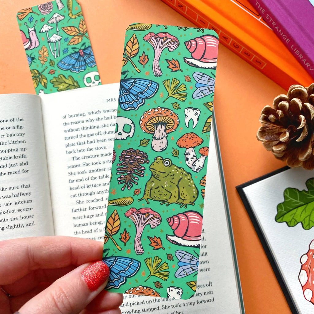 A green bookmark with a pattern of mushrooms, leaves, pinecones, and a large toad illustration. The background features an open book on an orange surface.