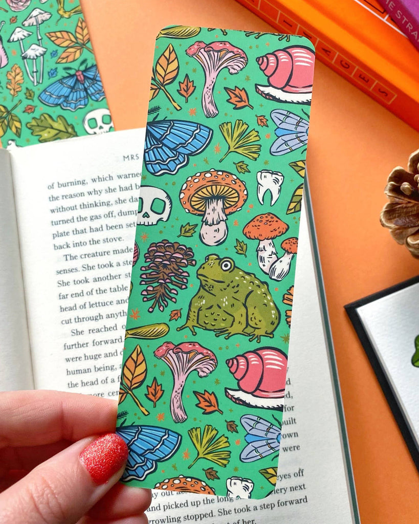 A green bookmark with a pattern of mushrooms, leaves, pinecones, and a large toad illustration. The background features an open book on an orange surface.