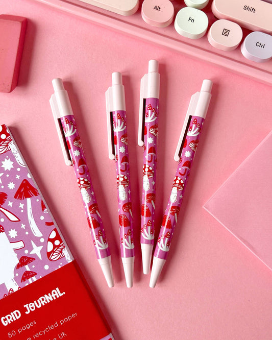 Four Rebel Romance pink mushroom pens are neatly arranged on a pink desk setup with pastel stationery. Designed by Stacey McEvoy Caunt, these pens feature a fun fungi-themed print for lovers of quirky and cute writing tools.