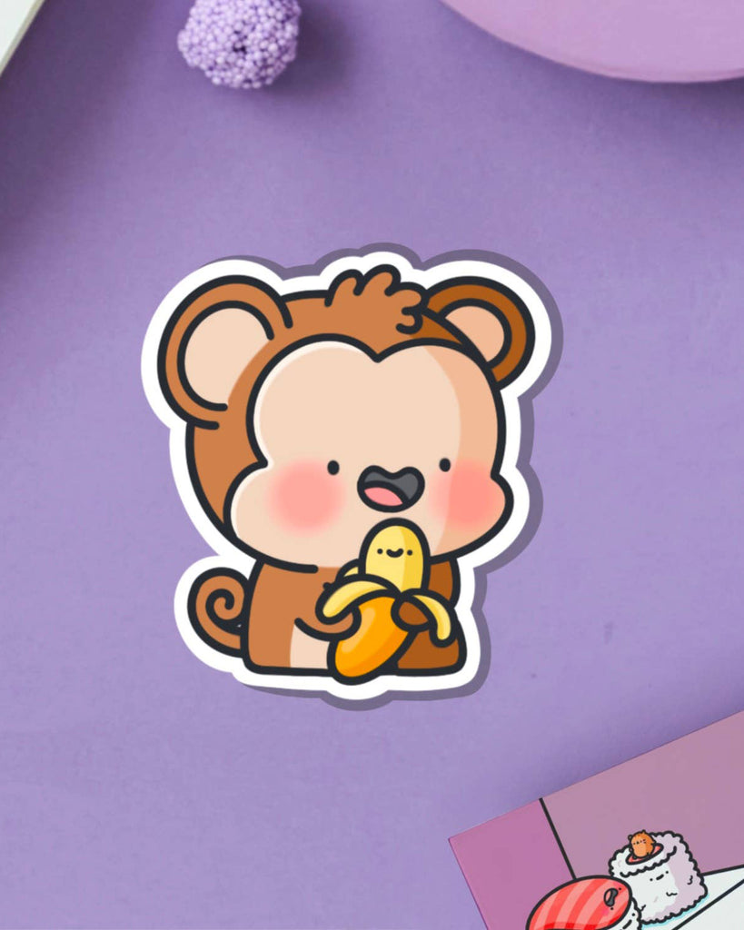 Adorable monkey vinyl sticker with a cheerful banana on a lilac purple background. A playful and colourful design by Toastedink, stocked by UK indie brand Rebel Romance.
