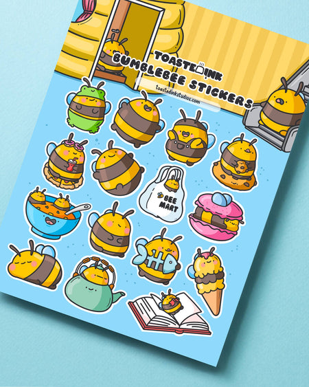 Close-up of the Toastedink Bumble Bee Sticker Sheet showcasing charming bumblebee illustrations on a soft blue background, placed on a pastel surface. Designs include bees dressed up in costumes like a chef, frog, soup bowl, and more, with bright and cheerful colours for craft and planner use.