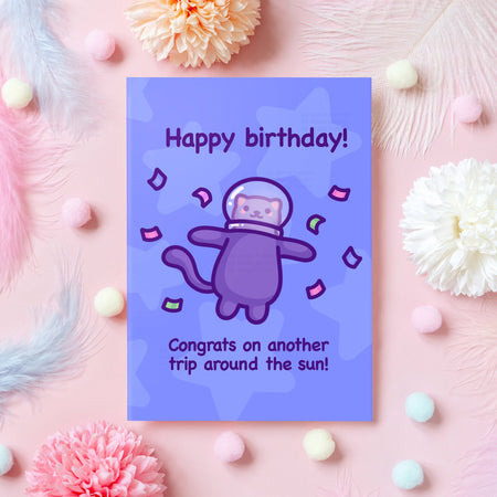 Cute space-themed birthday card by Mona Mauve for Rebel Romance, shown on a pink background with feathers, flowers, and pastel pom poms. The card features a purple cartoon cat in a space helmet surrounded by confetti, with the message “Happy birthday! Congrats on another trip around the sun!” A playful and colourful birthday card perfect for cat lovers and cosmic vibes.