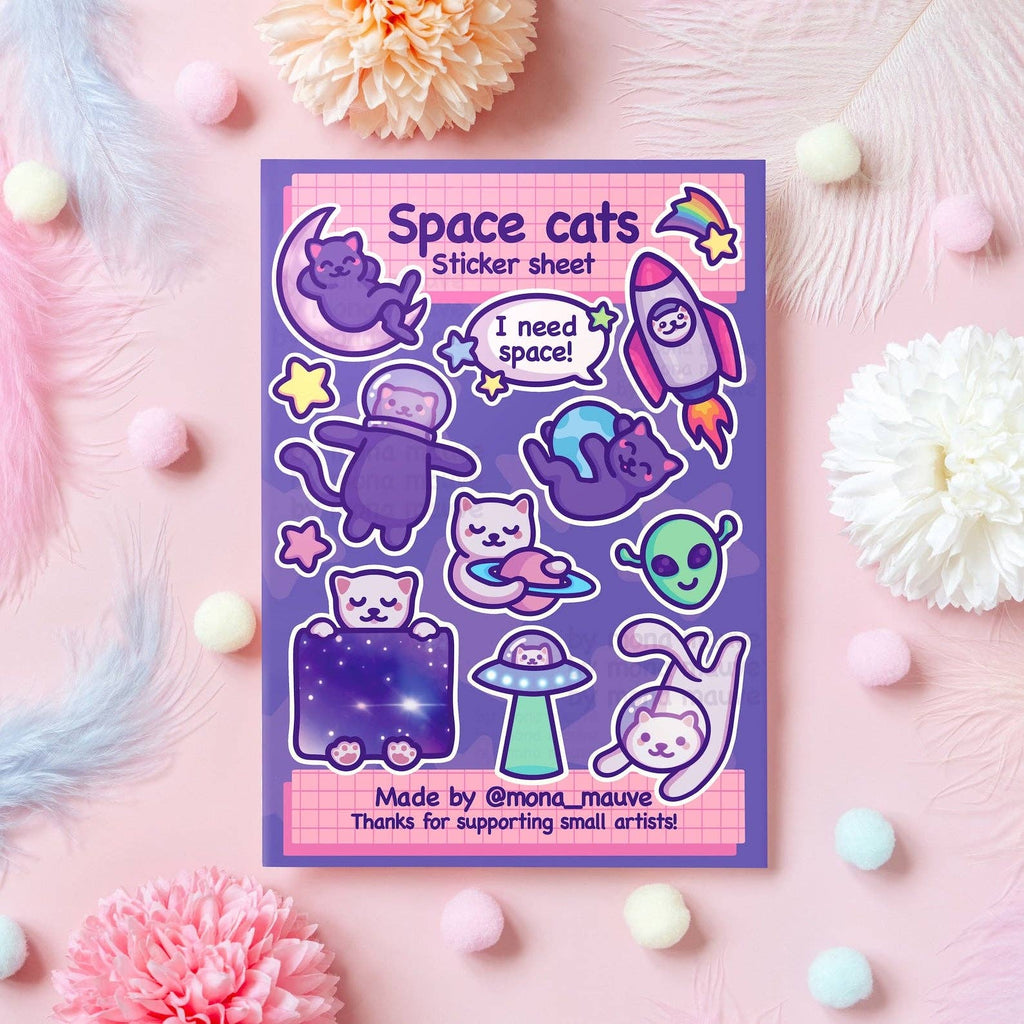 Flat lay of the Space Cats Sticker Sheet by Mona Mauve, featuring 13 waterproof vinyl stickers of adorable cartoon cats in space-themed designs like rockets, UFOs, planets, aliens, and the quote “I need space!” Displayed on a pink background with pastel pompoms and flowers. A perfect gift for cat lovers and space fans.