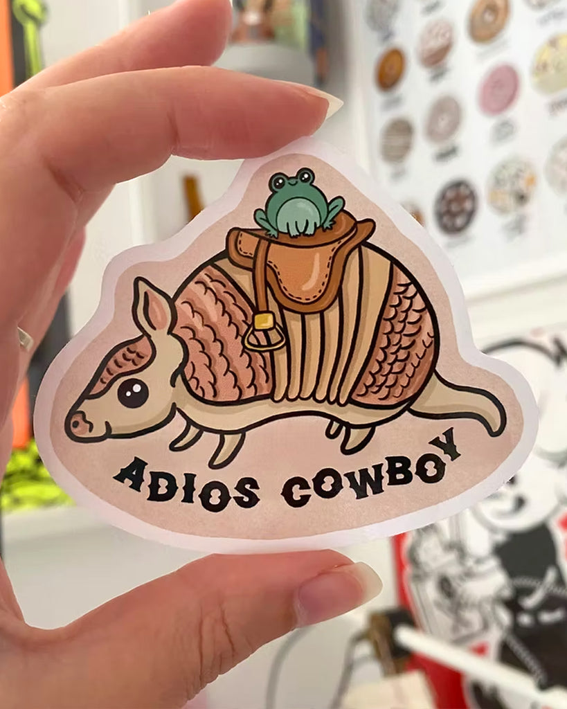 A vinyl sticker from Rebel Romance featuring a cartoon-style armadillo with a frog sitting in a saddle on its back. The armadillo is walking to the left, and the text “ADIOS COWBOY” appears in bold black letters below. Designed by Yasmin Cousins, this quirky, illustrated sticker is part of a range celebrating alternative and whimsical style. Ideal for decorating laptops, water bottles, or journals.