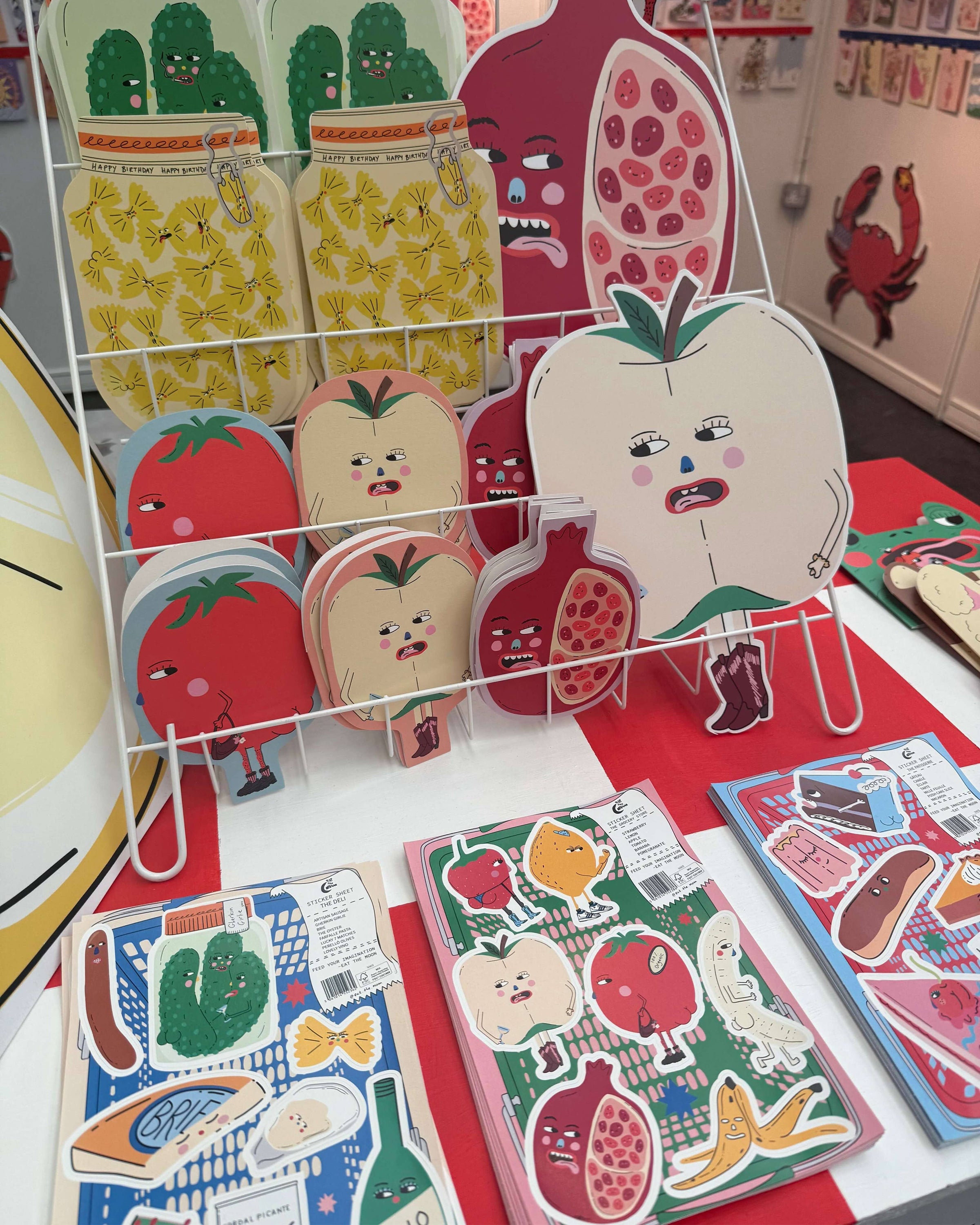 Close-up of Eat The Moon's quirky greeting cards and vinyl sticker sheets on display, designed by Eat The Moon. The bold and funny fruit and veg characters include tomatoes, pomegranates, pineapples, and more, with expressive faces and cheeky attitudes. Front and centre is the Patisserie A5 Vinyl Sticker Sheet, featuring cartoon food characters in bright colours. Display from a small UK business embracing alternative, millennial-friendly aesthetics and gifts.
