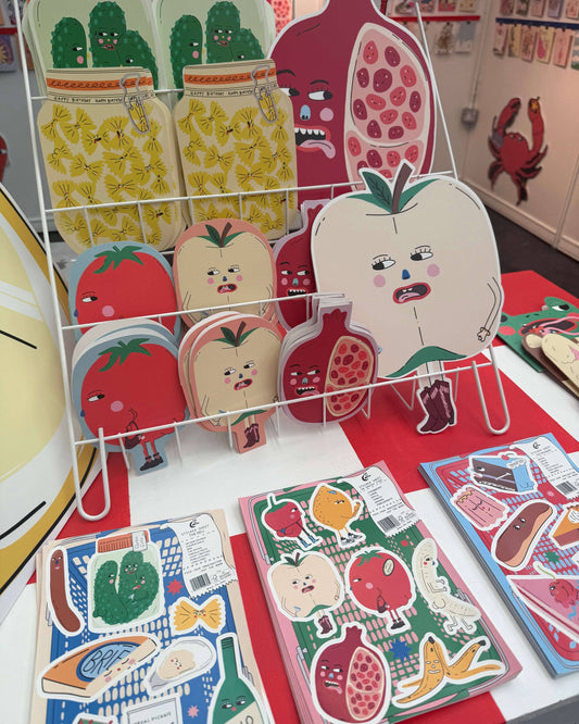 Close-up of Eat The Moon's quirky greeting cards and vinyl sticker sheets on display, designed by Eat The Moon. The bold and funny fruit and veg characters include tomatoes, pomegranates, pineapples, and more, with expressive faces and cheeky attitudes. Front and centre is the Patisserie A5 Vinyl Sticker Sheet, featuring cartoon food characters in bright colours. Display from a small UK business embracing alternative, millennial-friendly aesthetics and gifts.