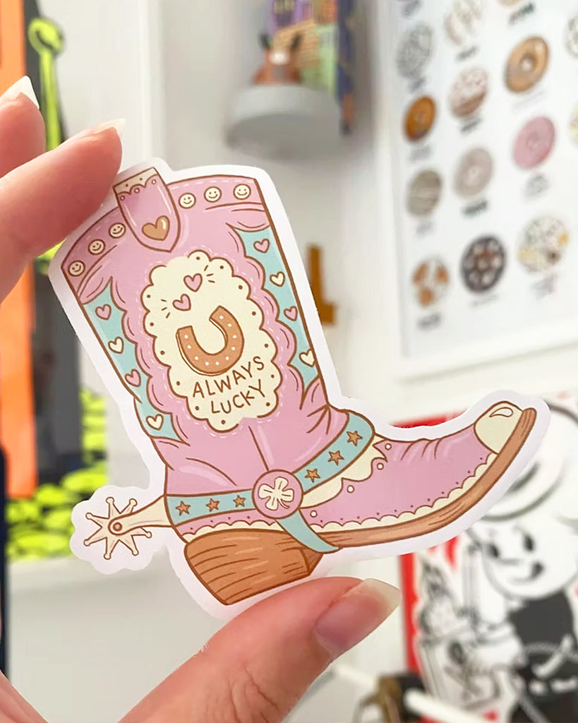 A hand holding a Rebel Romance vinyl sticker shaped like a pastel pink and blue cowboy boot with a horseshoe reading "Always Lucky" in the centre. Designed by Yasmin Cousins, this cute Western-themed sticker features heart, star and smiley face details. Perfect for decorating laptops, water bottles or journals. A unique accessory from UK brand Rebel Romance, known for its alternative, millennial-friendly designs.