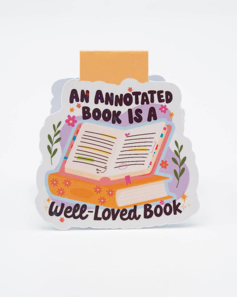 Front view of the An Annotated Book is a Well-Loved Book Magnetic Bookmark from Rebel Romance. Features a pastel illustration of an open book with colourful notes and underlining, sitting atop a stack of books with floral details. A sweet and aesthetic stationery item for readers who love making their books personal and expressive.