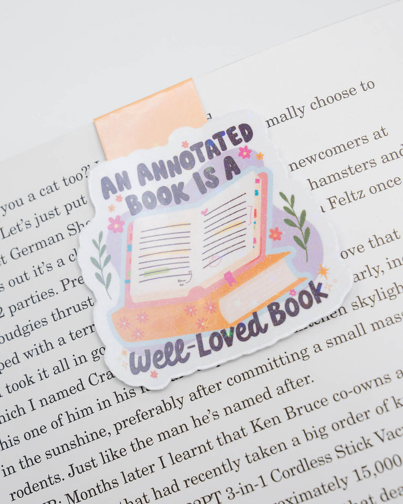 Magnetic bookmark clipped to a book page, featuring pastel colours and the quote “An Annotated Book is a Well-Loved Book.” Illustrated with a marked-up open book, flowers, and soft sparkles. A perfect accessory for readers who love to annotate, highlight, and make books their own. Sold by Rebel Romance, a UK brand for quirky, book-loving millennials.
