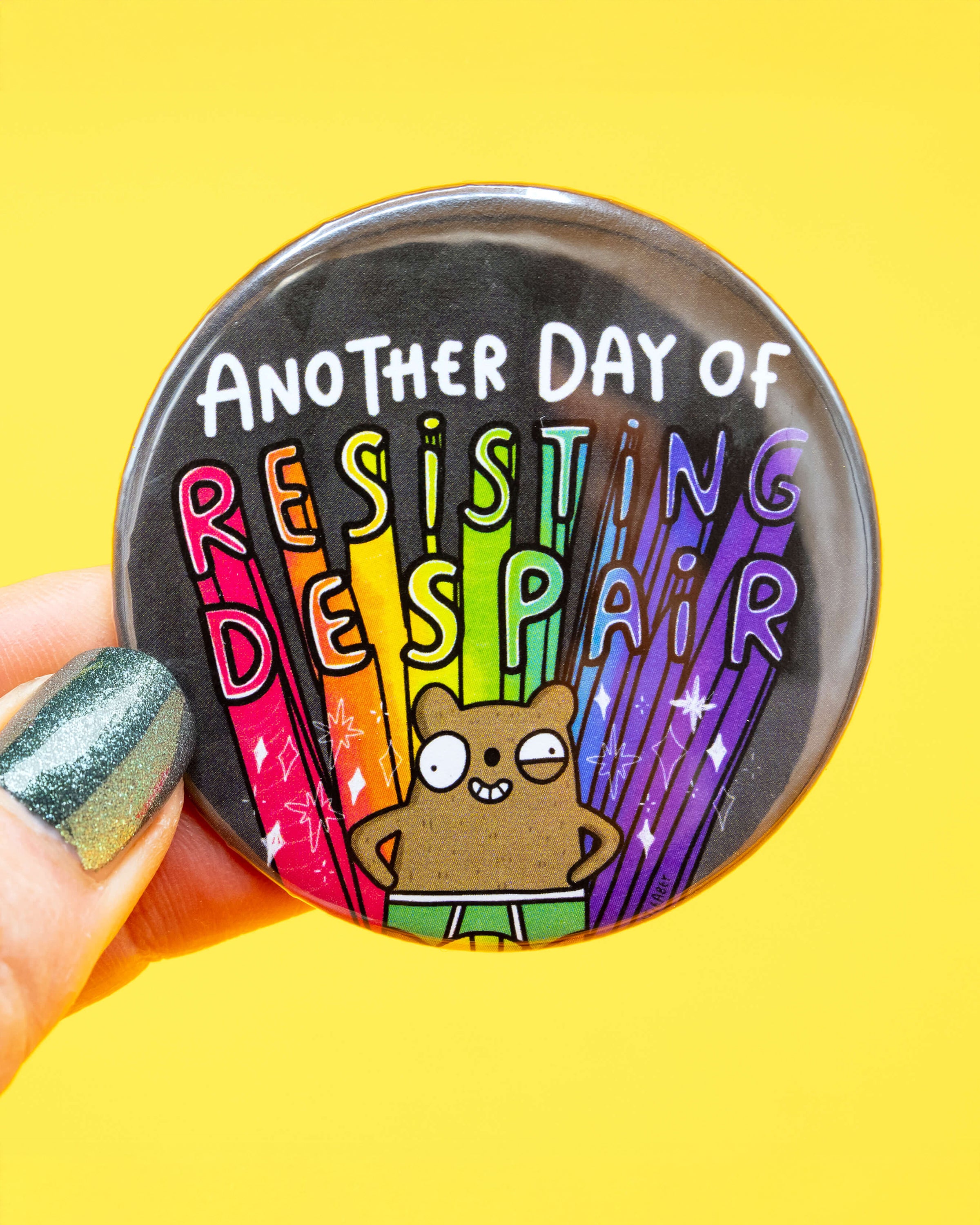 Rebel Romance 'Resisting Despair Bear Badge' held in someone's fingers against a bright yellow background. The design shows a happy cartoon bear in rainbow shorts with the words “Another Day of Resisting Despair” in rainbow lettering. A cheerful and empowering accessory from Katie Abey.