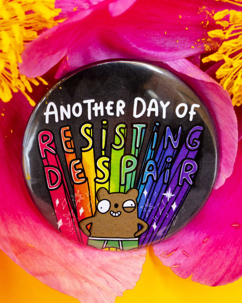 Close-up of the Rebel Romance 'Resisting Despair Bear Badge' nestled inside a vibrant pink flower. The circular badge features a cartoon bear in rainbow pants standing proudly with the phrase "Another Day of Resisting Despair" in bold rainbow letters. A colourful and quirky motivational badge by Katie Abey.
