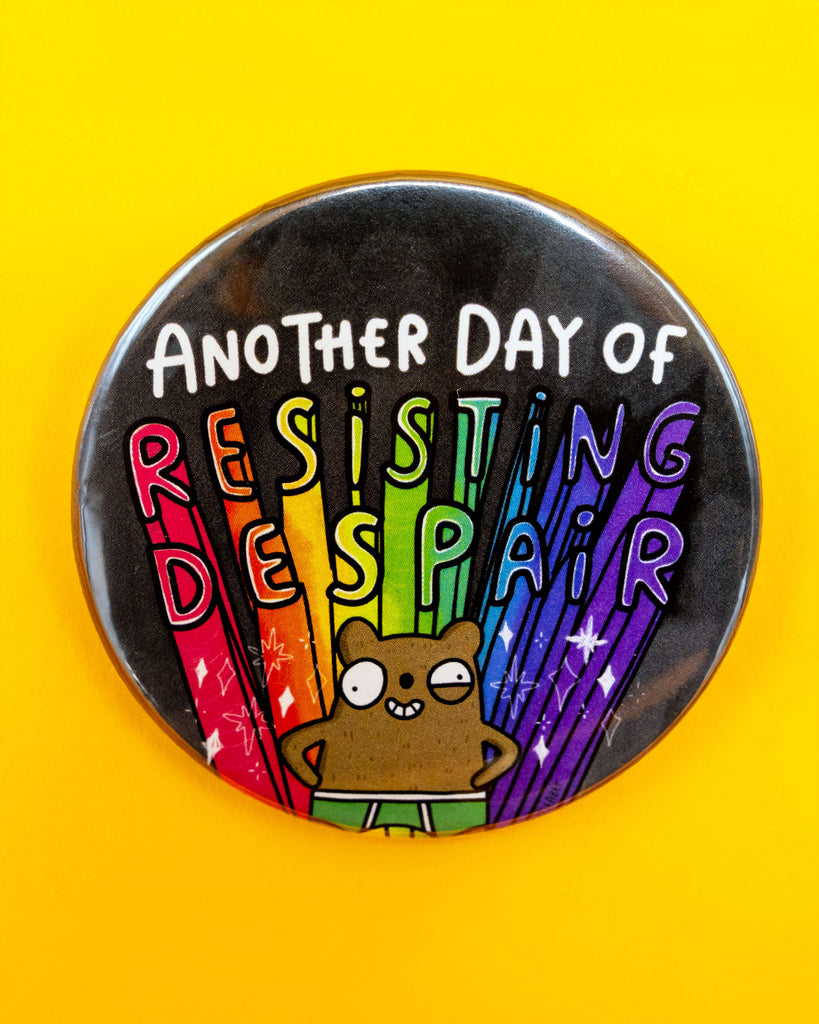 Flat-lay photo of the Rebel Romance 'Resisting Despair Bear Badge' by Katie Abey on a yellow background. The badge shows a cute cartoon bear smiling under rainbow beams and the words “Another Day of Resisting Despair,” symbolising resilience and queer joy.