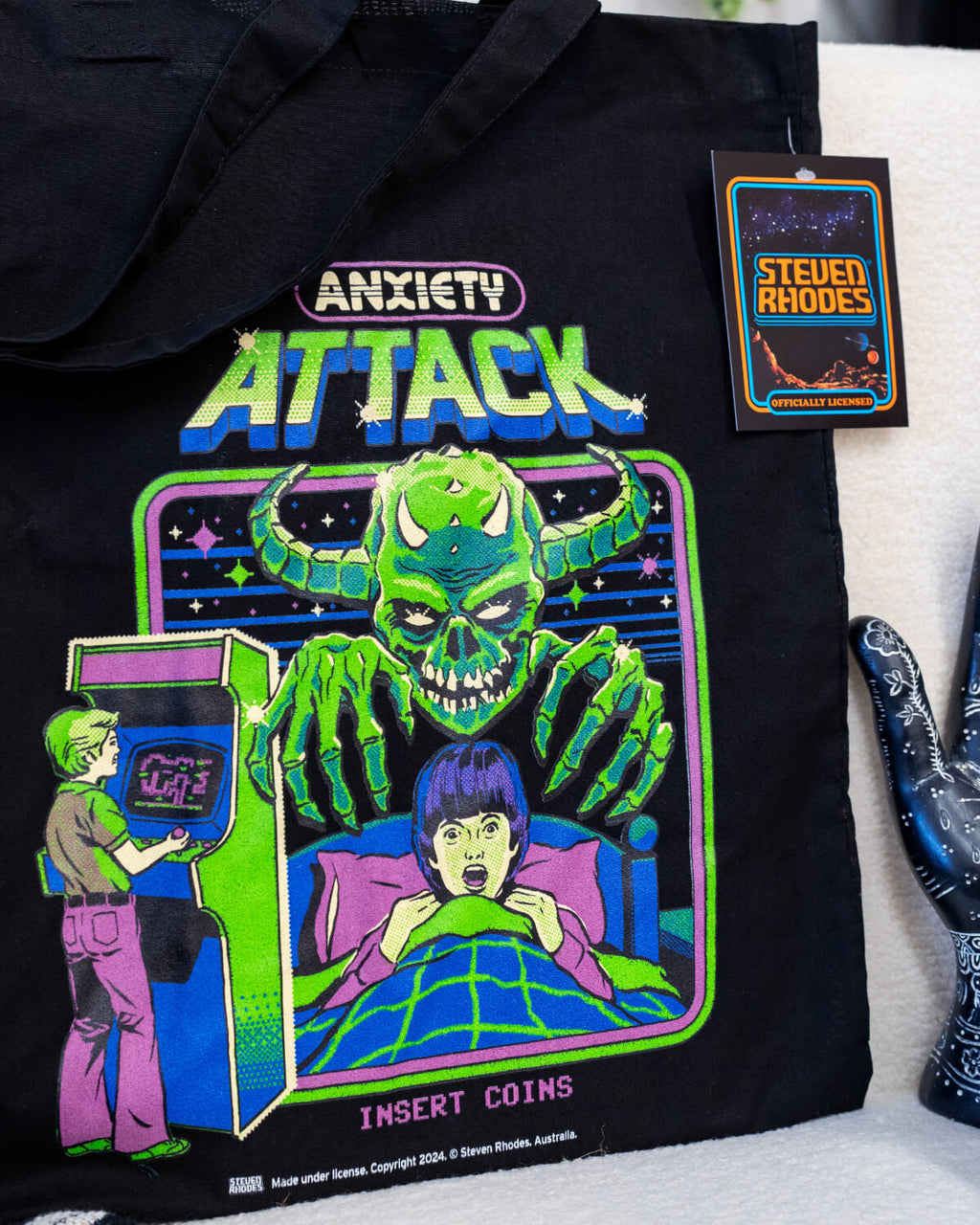 A black tote bag featuring a Steven Rhodes design titled “Anxiety Attack.” The artwork depicts a green monster with glowing eyes and sharp claws looming over a person in bed, while another person plays an arcade game in the background. Neon text reads “Anxiety Attack” and “Insert Coins.” The vibrant, retro style contrasts with the dark background, adding a playful yet eerie vibe.