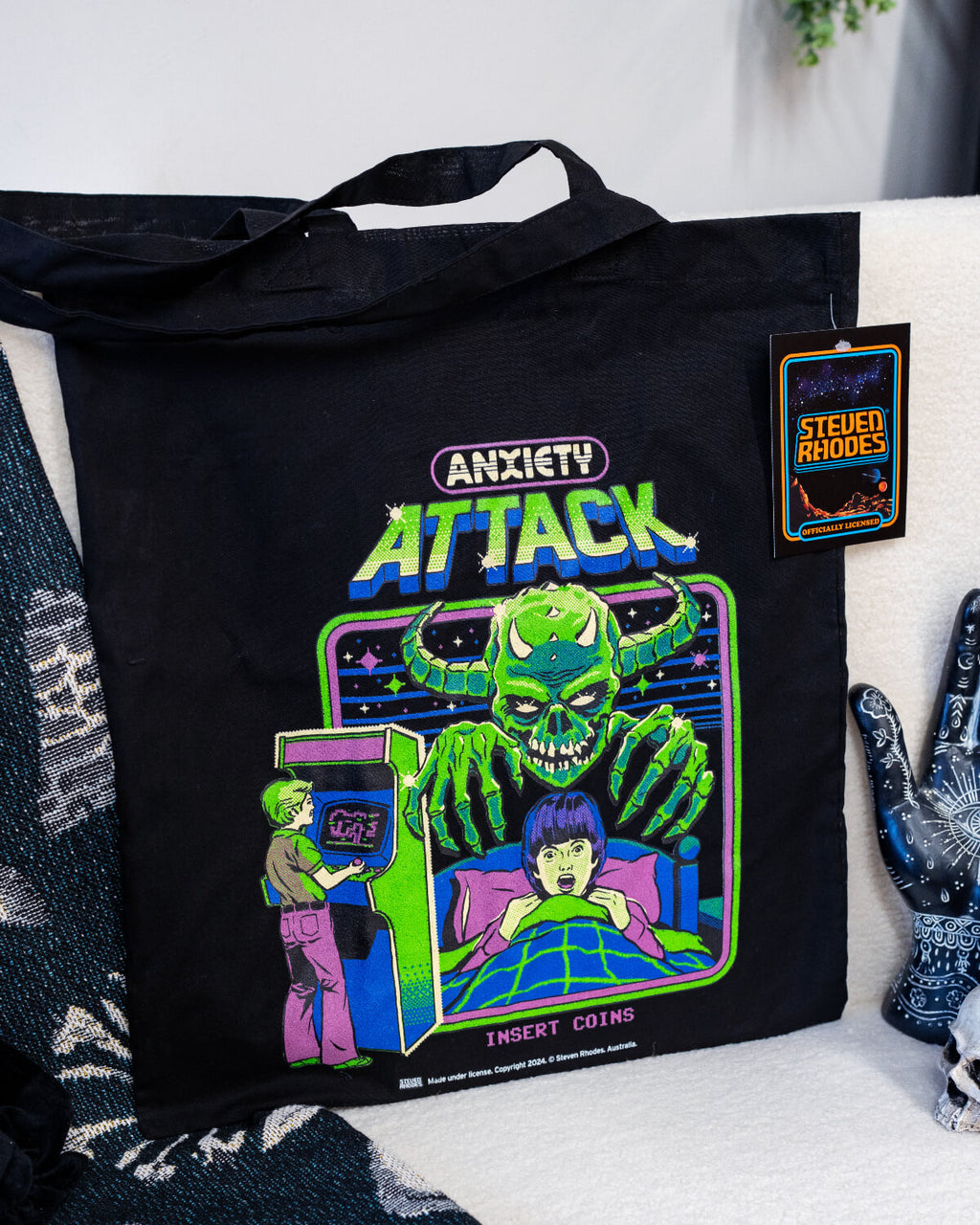 A black tote bag featuring a Steven Rhodes design titled “Anxiety Attack.” The artwork depicts a green monster with glowing eyes and sharp claws looming over a person in bed, while another person plays an arcade game in the background. Neon text reads “Anxiety Attack” and “Insert Coins.” The vibrant, retro style contrasts with the dark background, adding a playful yet eerie vibe.