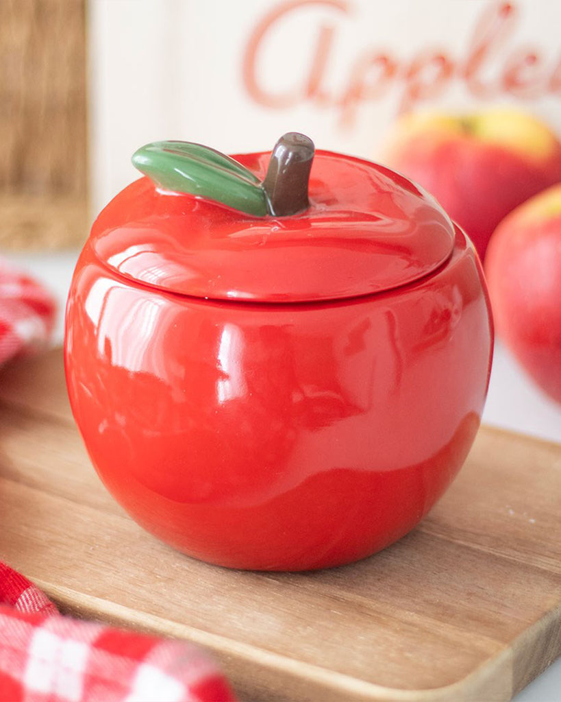 Red ceramic apple-shaped candle jar with the lid on, designed with a green leaf and brown stem on top, sitting on a wooden tray near red apples and a red gingham cloth.